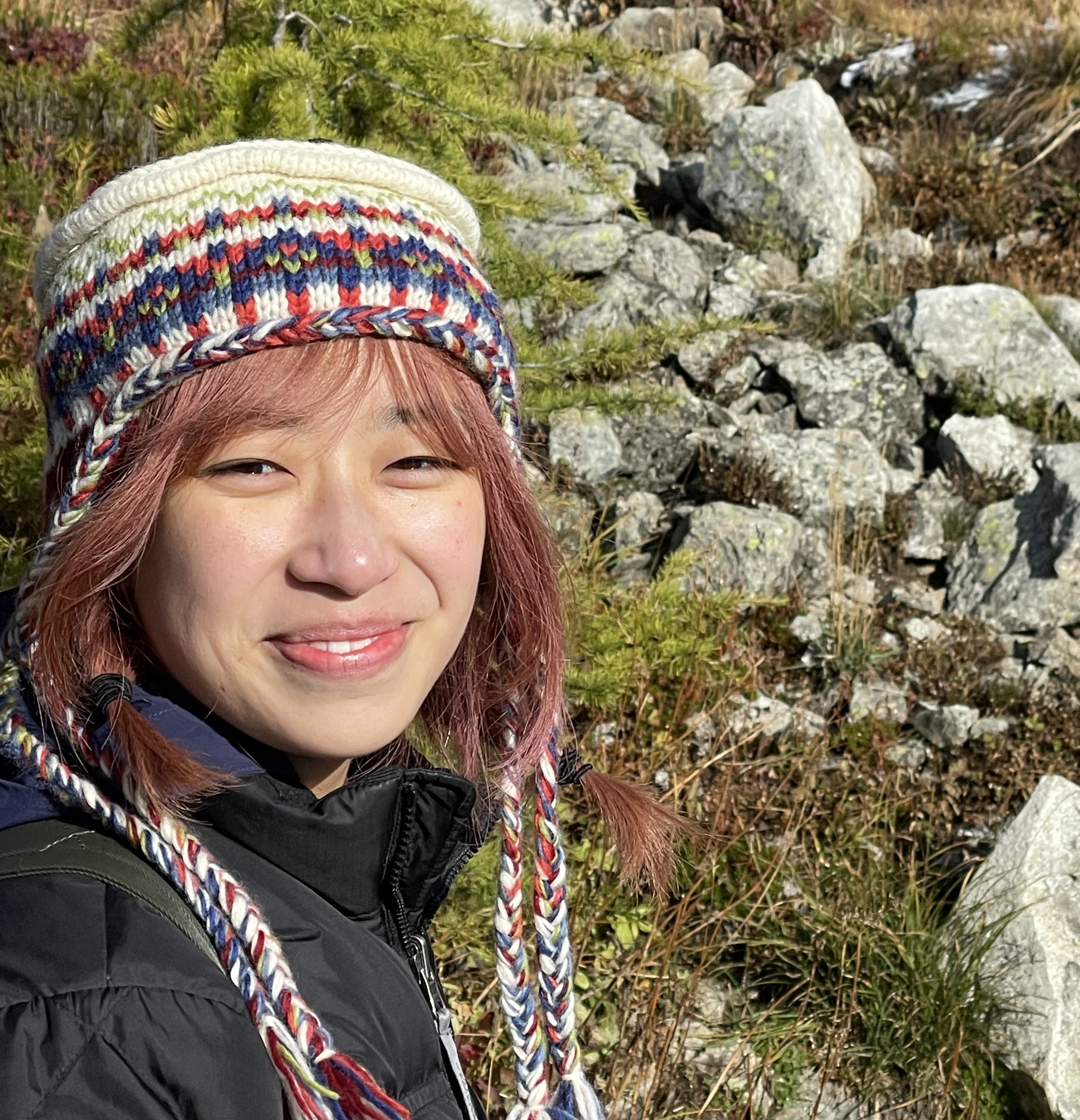 chinese-canadian woman with pink hair in braids and an icelandic wool hat smiling at the camera with a larch tree and rocks in the background